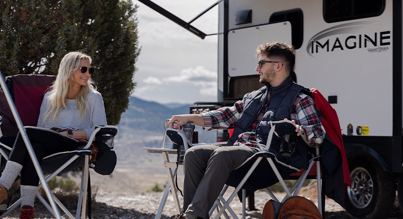 Couple sitting outdoors in front of a Grand Design Imagine travel trailer.